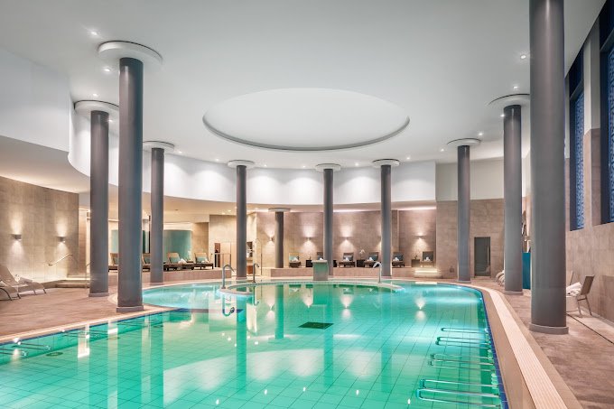 Indoor swimming pool area with clear water, surrounded by lounge chairs and seating areas, with tall columns and soft ambient lighting.