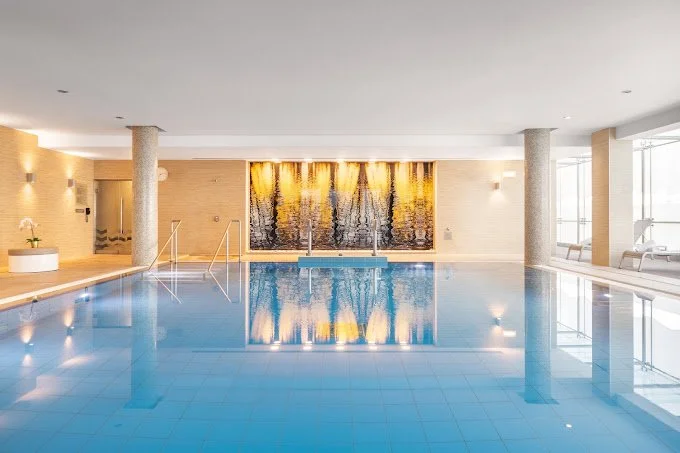 Indoor swimming pool with blue water, surrounded by beige walls, large windows, and modern decor.