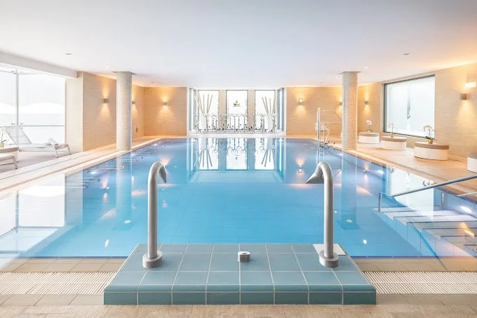 Indoor swimming pool with steps and handrails, lounge chairs, large windows, and soft lighting.