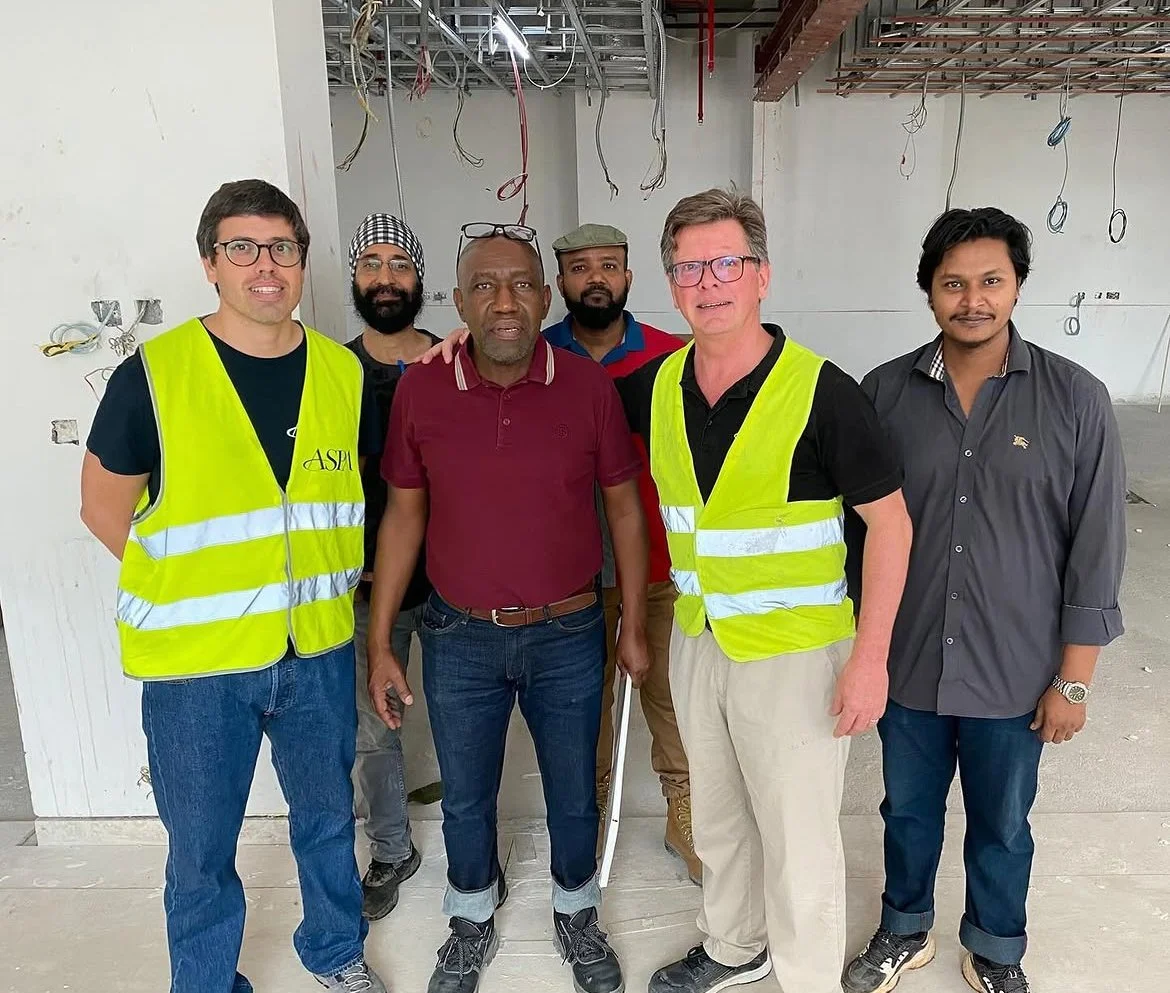 Group of six men standing inside a building under construction, some wearing safety vests, with exposed wires and unfinished walls behind them.