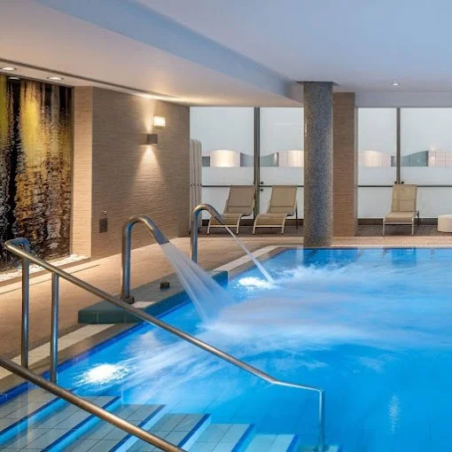 Indoor heated swimming pool with waterfalls, lounge chairs, and large windows in a modern spa or hotel.