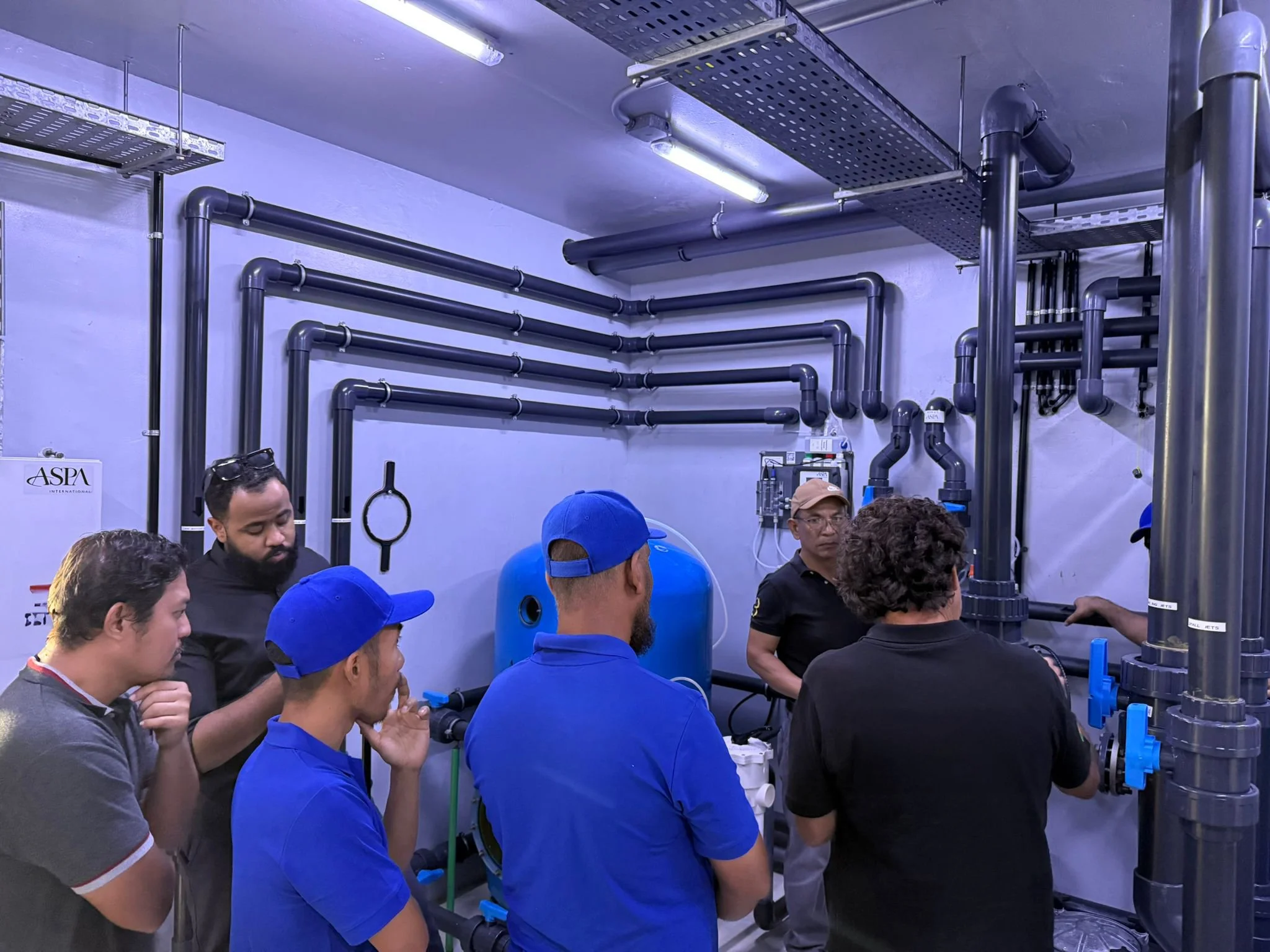 Group of people wearing blue caps and black shirts gathered in an industrial room with pipes, metal shelves, and equipment, listening to a person explaining.