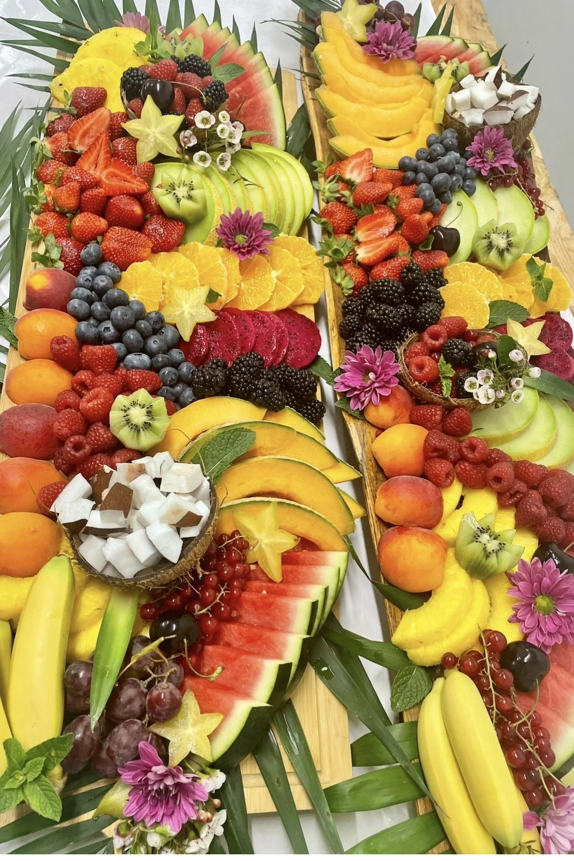 Plat de fruits frais découpés, comprenant pastèque, kiwi, fraises, mûres, myrtilles, orange, melon, bananes, pommes, raisins, et garnitures florales.