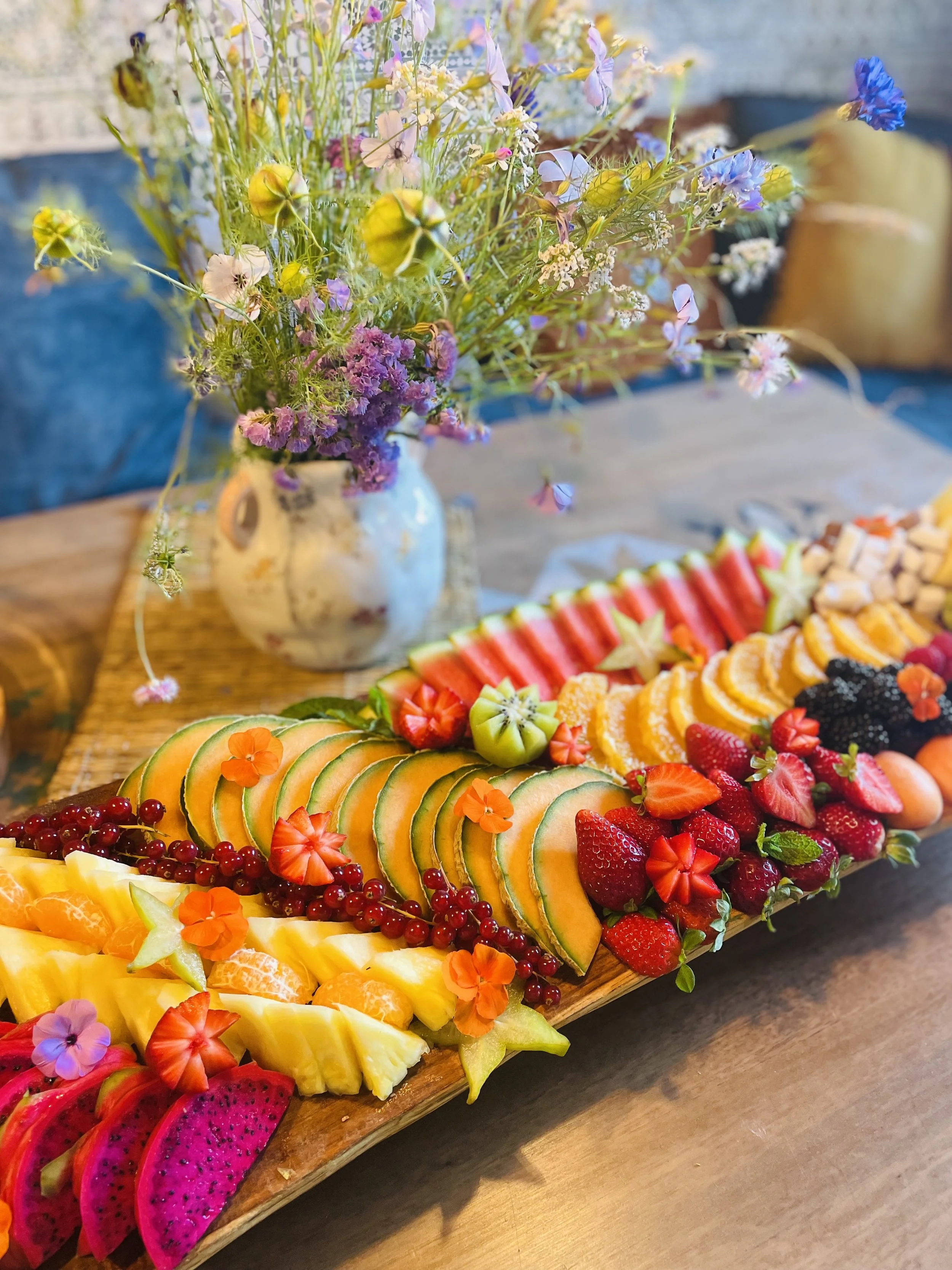 Plateau de fruits frais tranchés, comprenant pastèque, kiwi, fraises, ananas, mangue, et autres fruits, décoré avec des fleurs comestibles.