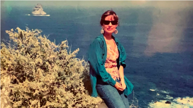 A woman wearing sunglasses and a teal jacket sitting on rocks near the ocean, with a boat in the background.