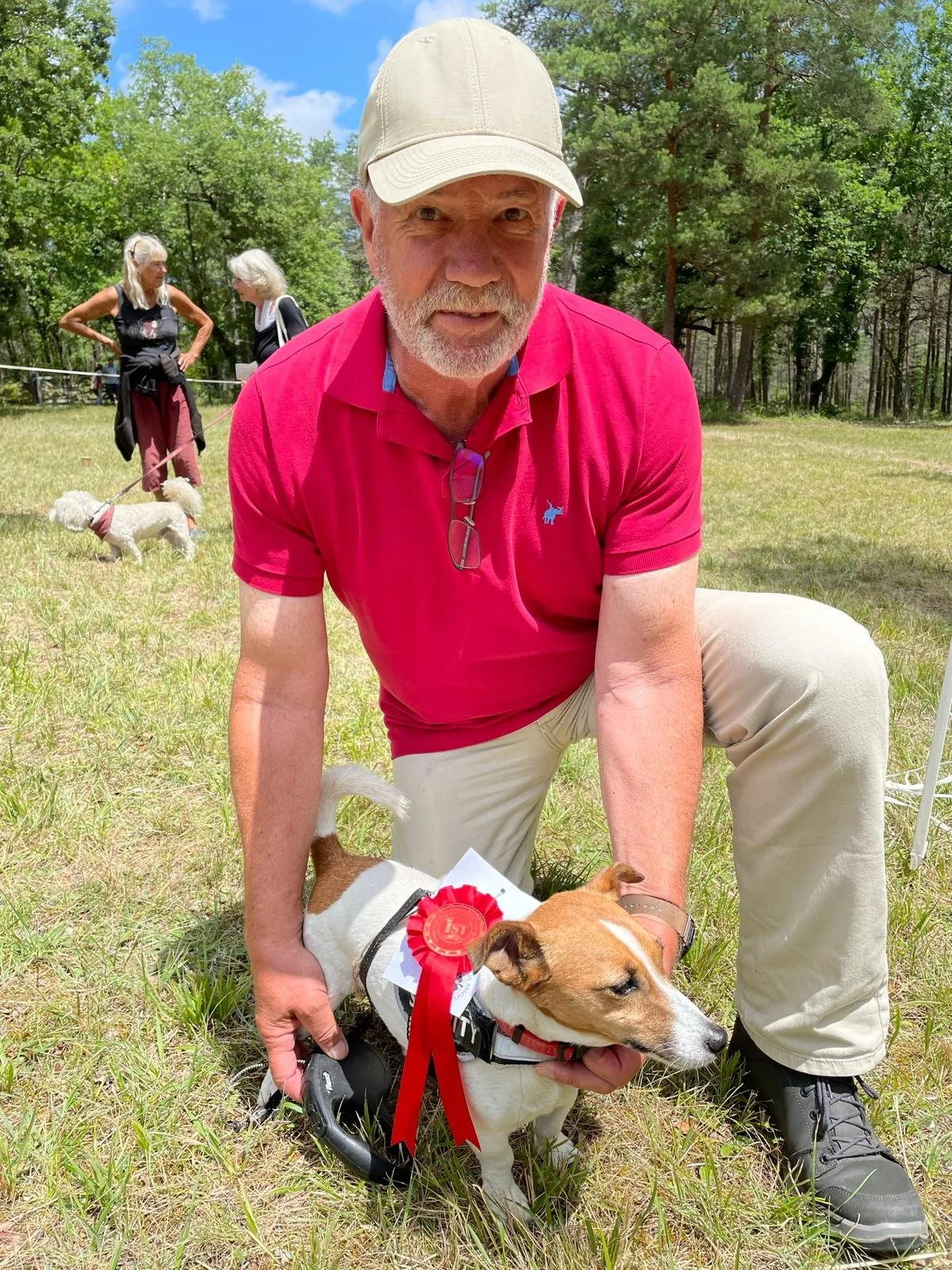 A man squats on grass holding a dog with a red rosette ribbon. In the background, two women are walking dogs on leashes in a park with trees.