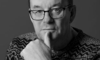 Black and white portrait of a man wearing glasses and a patterned sweater, with his hand touching his chin in a thoughtful pose.