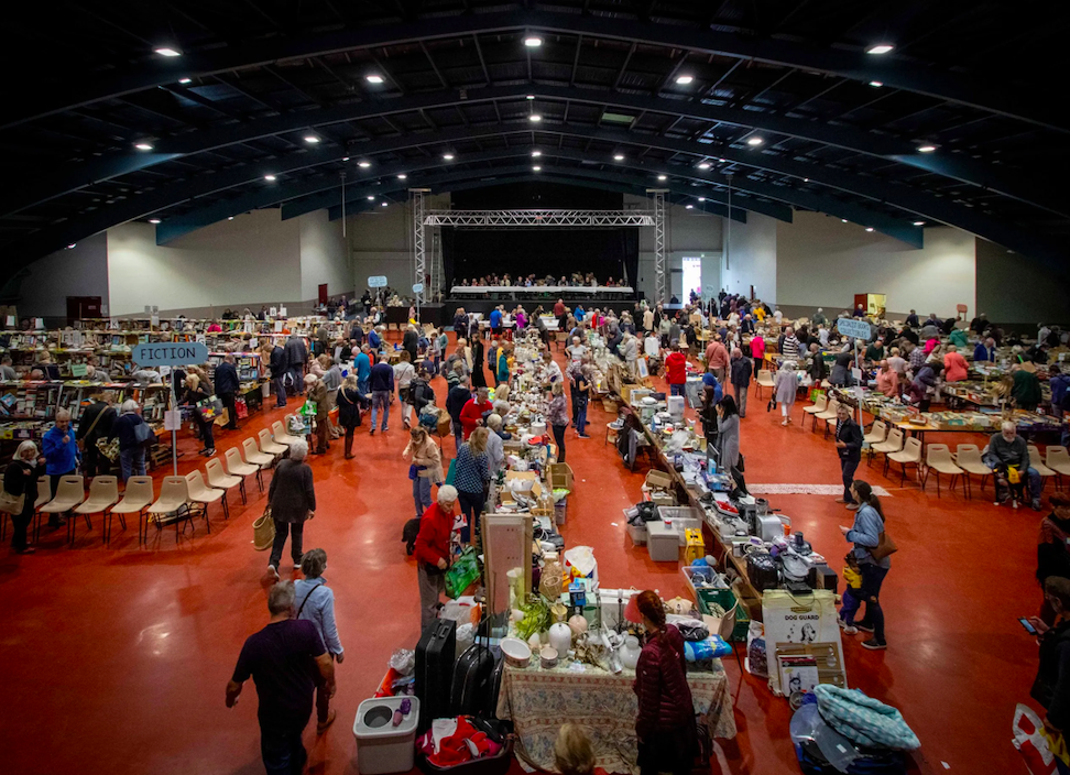 Indoor flea market with multiple tables filled with various items, people browsing, and a stage at the back.