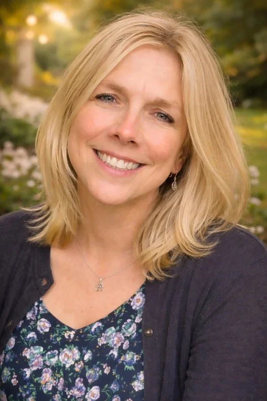 A woman with blonde hair smiling outdoors with blurred greenery and flowers in the background.