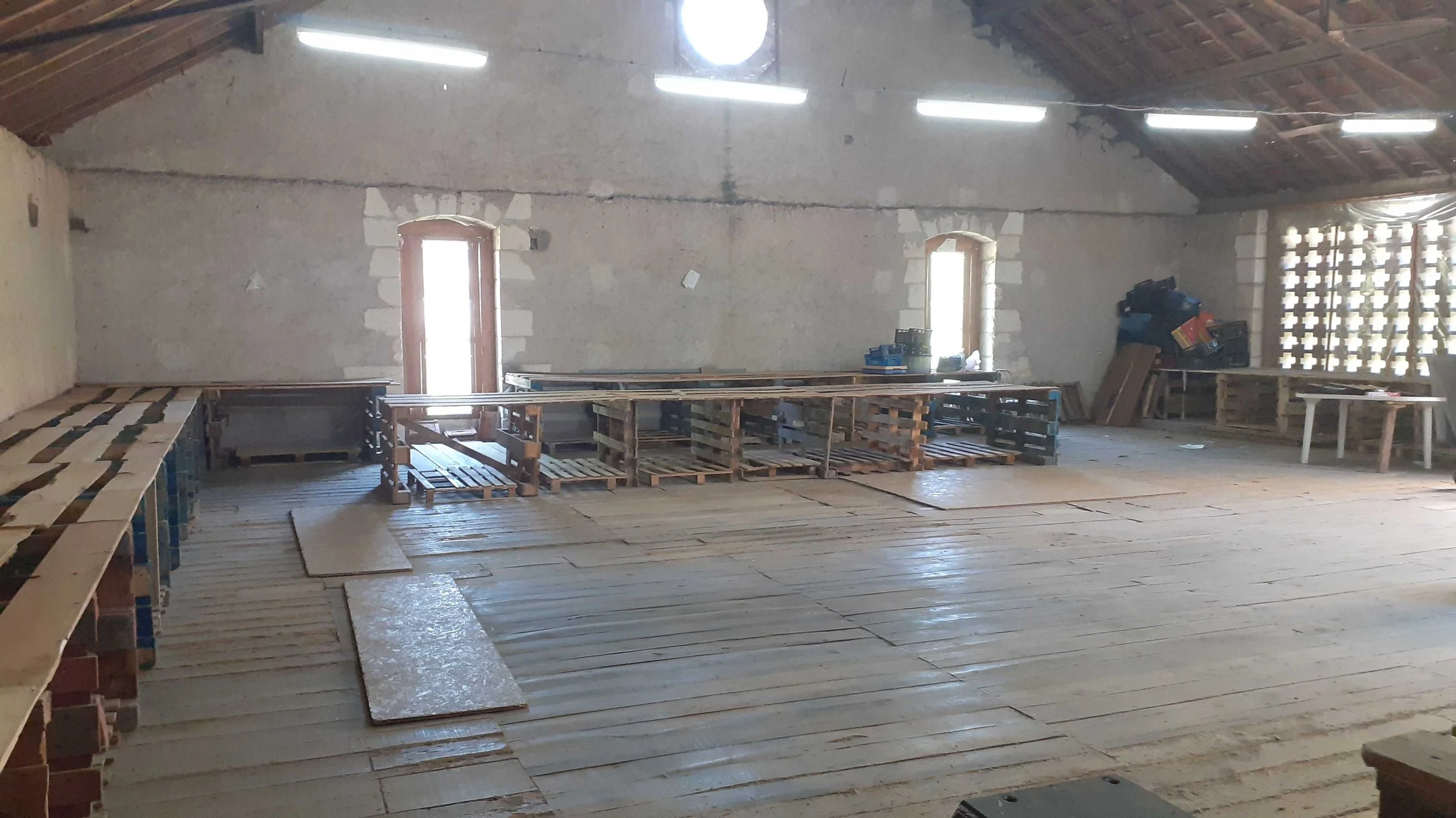 Interior of an unfinished building with wooden flooring, construction materials, and pallets, with high ceilings, small windows, and exposed brick walls.