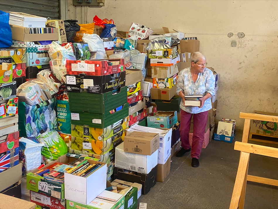 A woman standing among a large pile of boxes and bags filled with various items, in a warehouse or storage room setting.