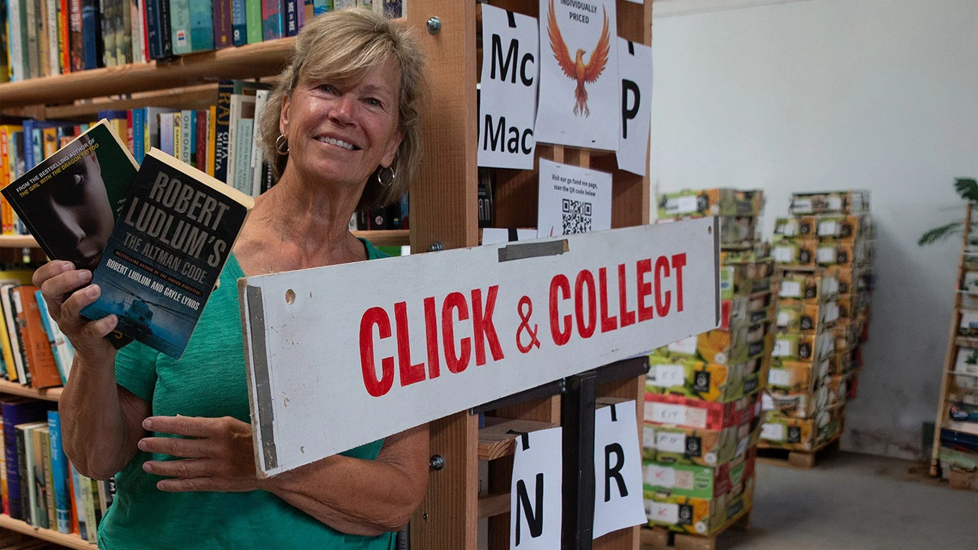 A woman smiling in a bookstore, holding two books and standing next to a sign that says "Click & Collect" with shelves of books in the background.