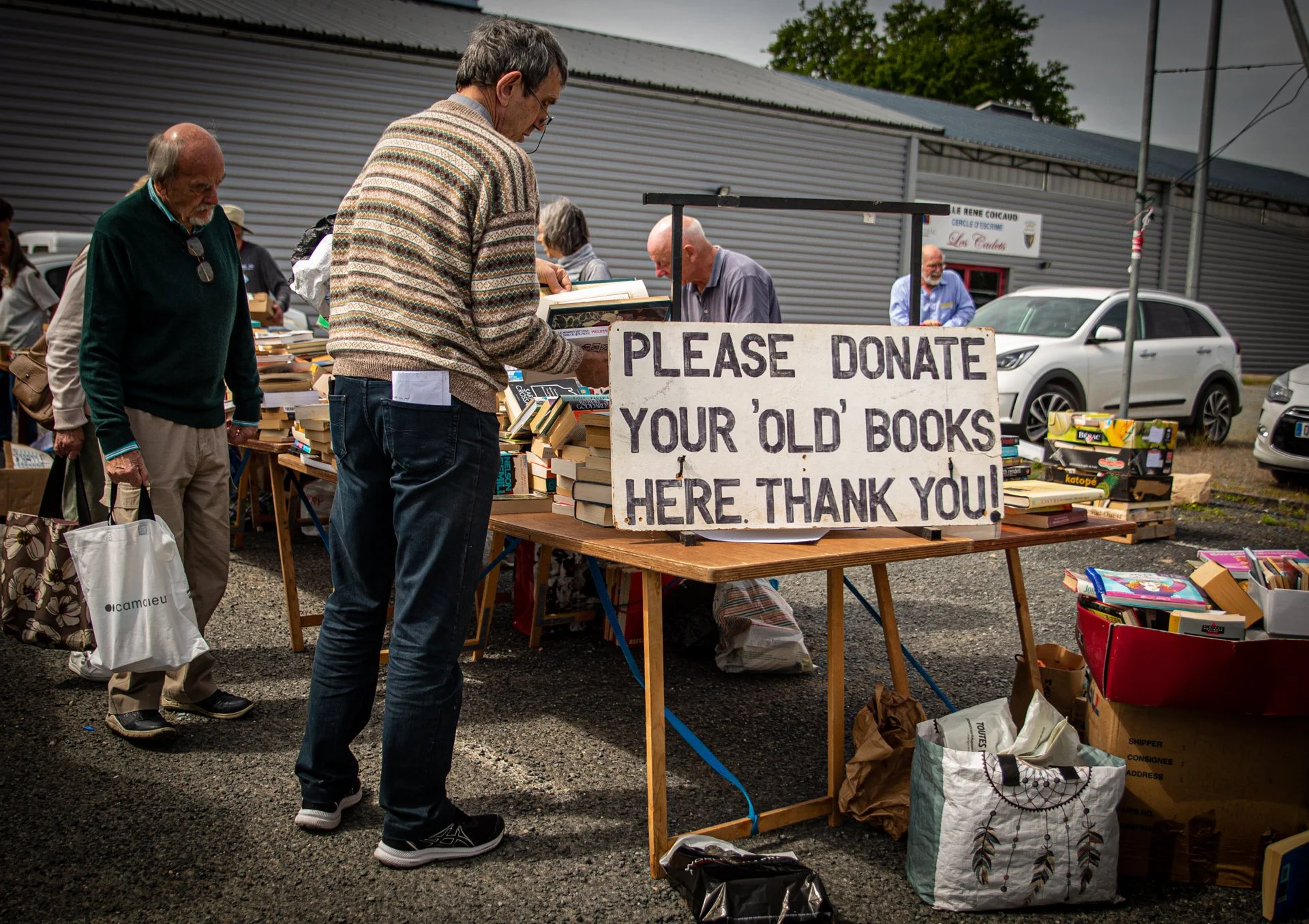 People browsing used books at an outdoor book sale. There is a sign reading, 'Please donate your old books. Here thank you!'