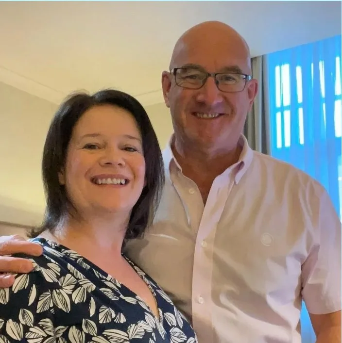A smiling woman with dark hair and a patterned blouse standing next to a smiling man with glasses and a light pink shirt in a well-lit room with curtains and blue lighting in the background.