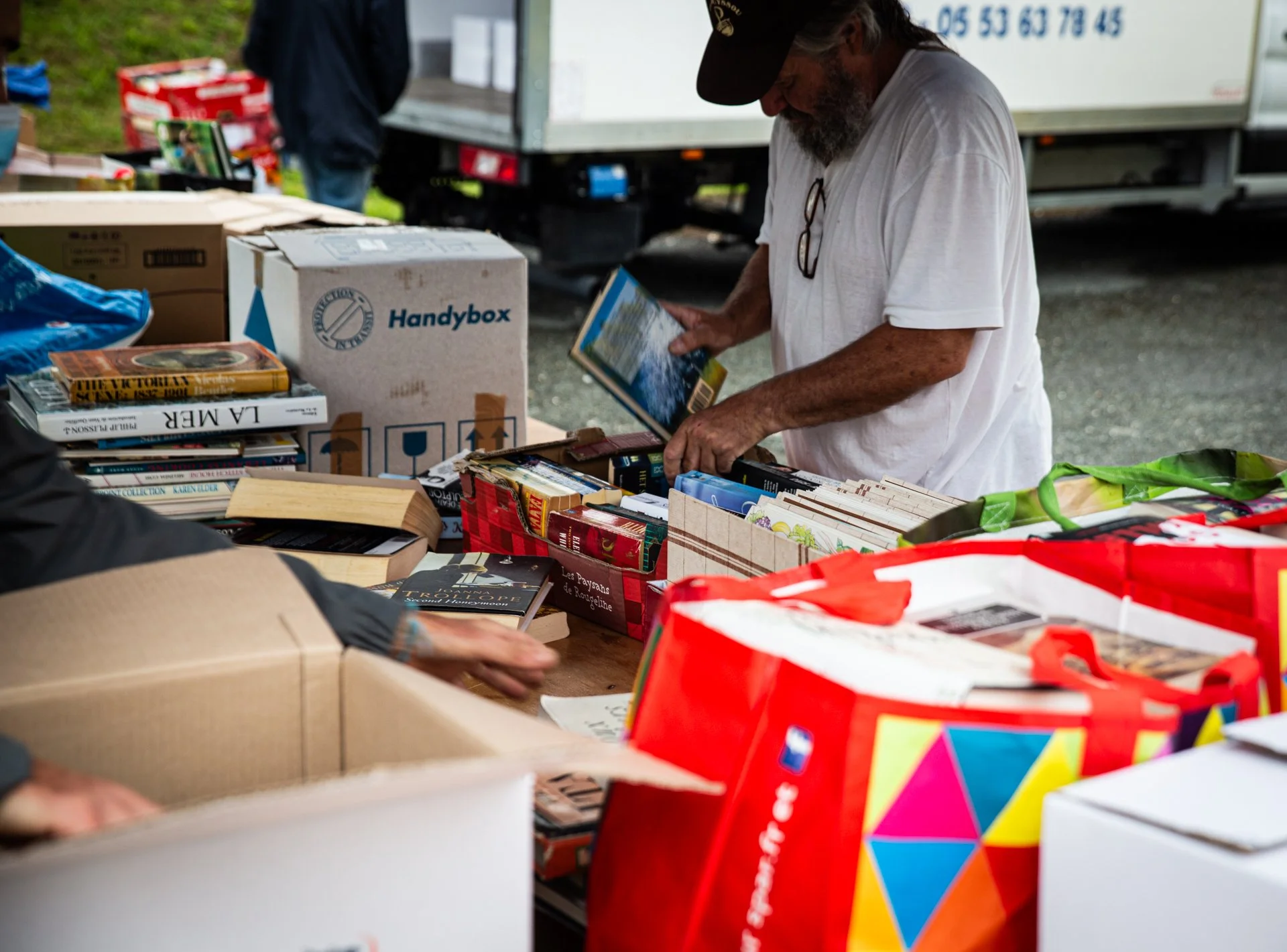 A man with a gray beard wearing a white shirt and a black cap browsing books at an outdoor sale table. The table is filled with books and boxes, and there are other people and a truck in the background.