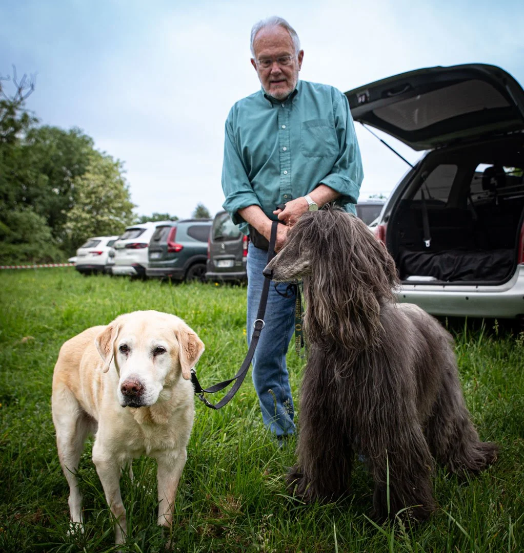 Older man standing outdoors with two dogs, a Labrador Retriever and a large long-haired dog, near an open car trunk, with parked cars and greenery in the background.