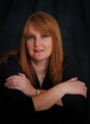 Portrait of a woman with red hair, wearing a black top and jewelry, resting her arms on a surface in front of her.