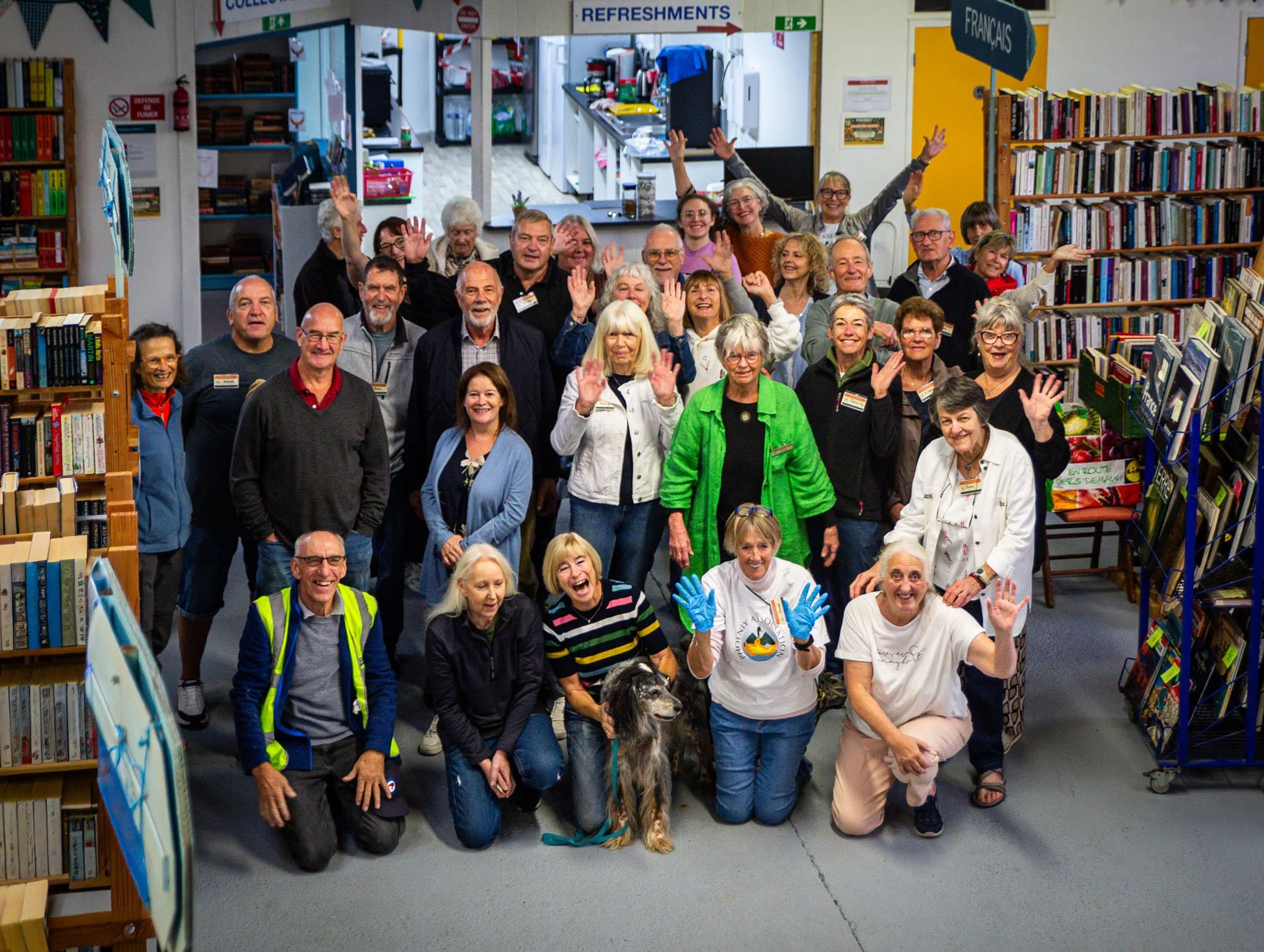 A group of adults and a dog in a bookstore or library, smiling and waving at the camera. The background features bookshelves, signs for refreshments and French books, and a kitchen area.