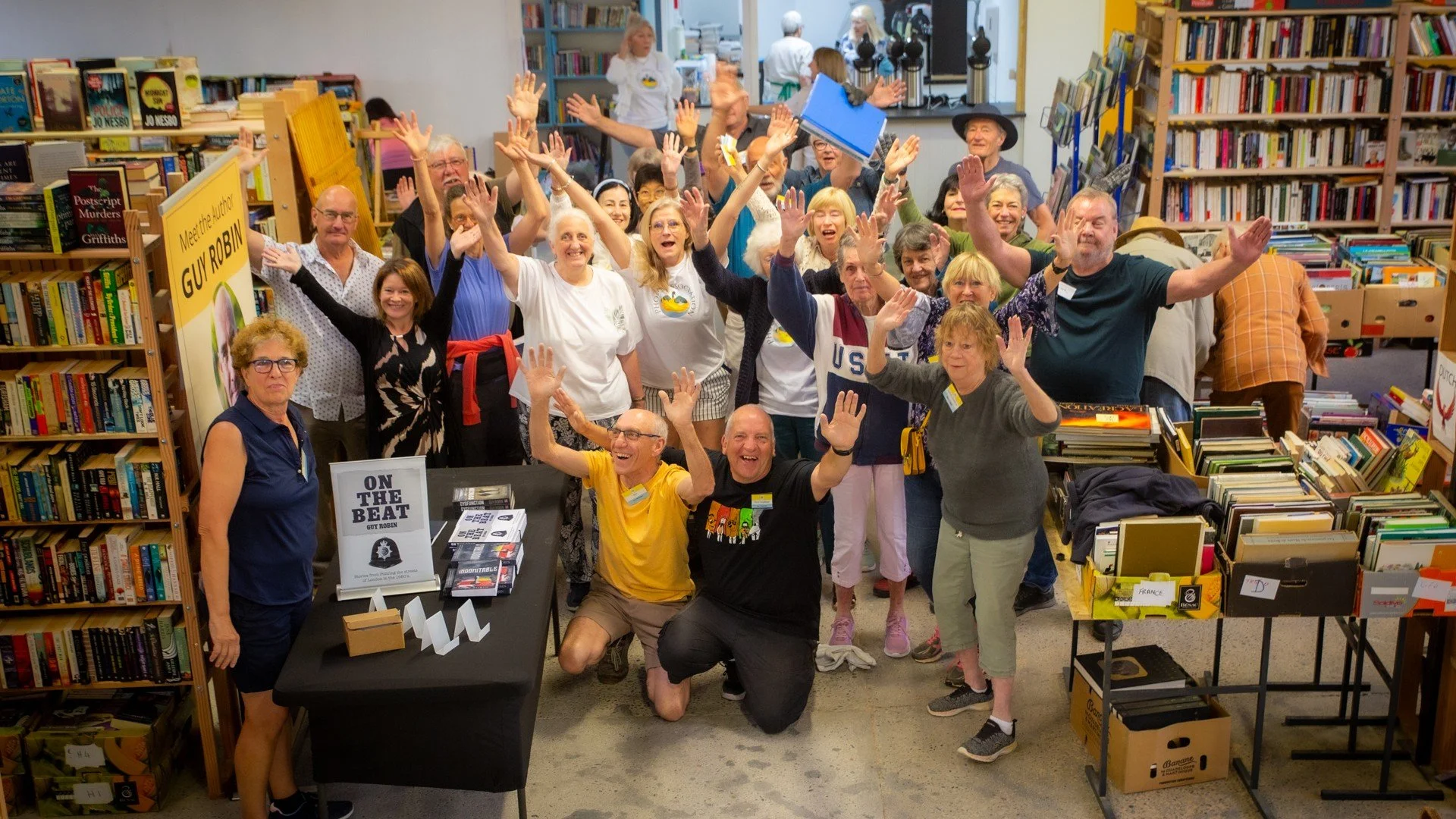 A group of people at a book fair or event inside a bookstore, smiling, raising their hands, and posing for the photo. Bookshelves and tables with books surround them.