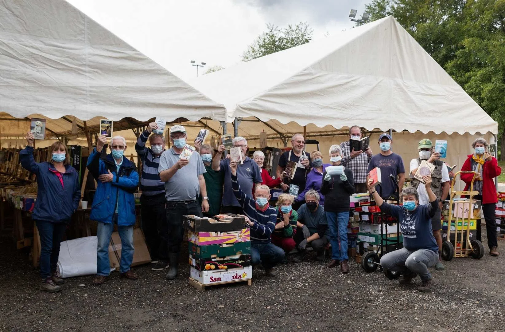 Group of people wearing masks at a book sale or market under a large white tent, holding books and smiling for the photo.