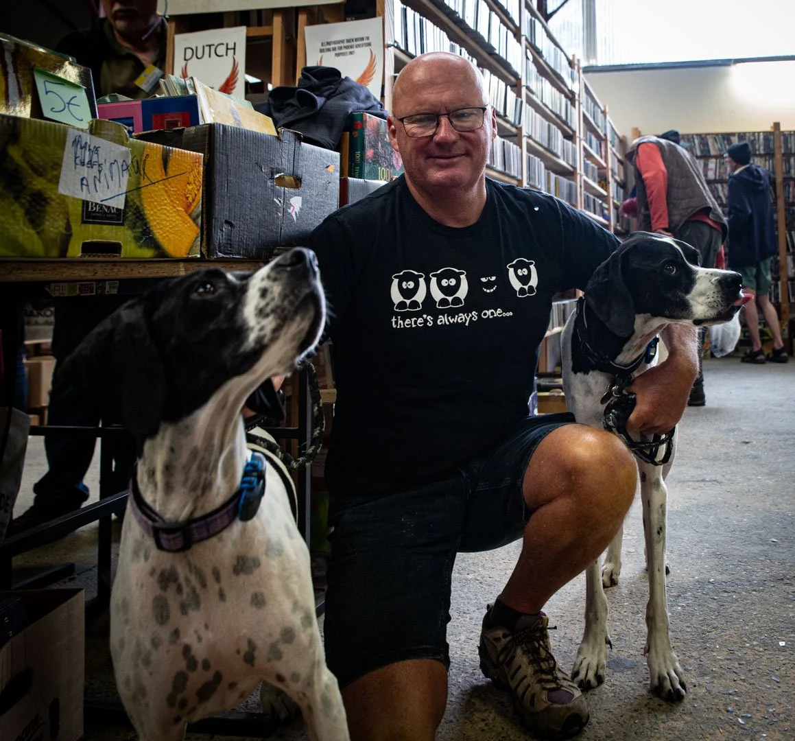 A man with glasses kneeling inside a bookstore, holding two black and white dogs. The man is wearing a black t-shirt with cartoon sheep and the text 'there's always one.' In the background, there are shelves filled with books and other people browsing.
