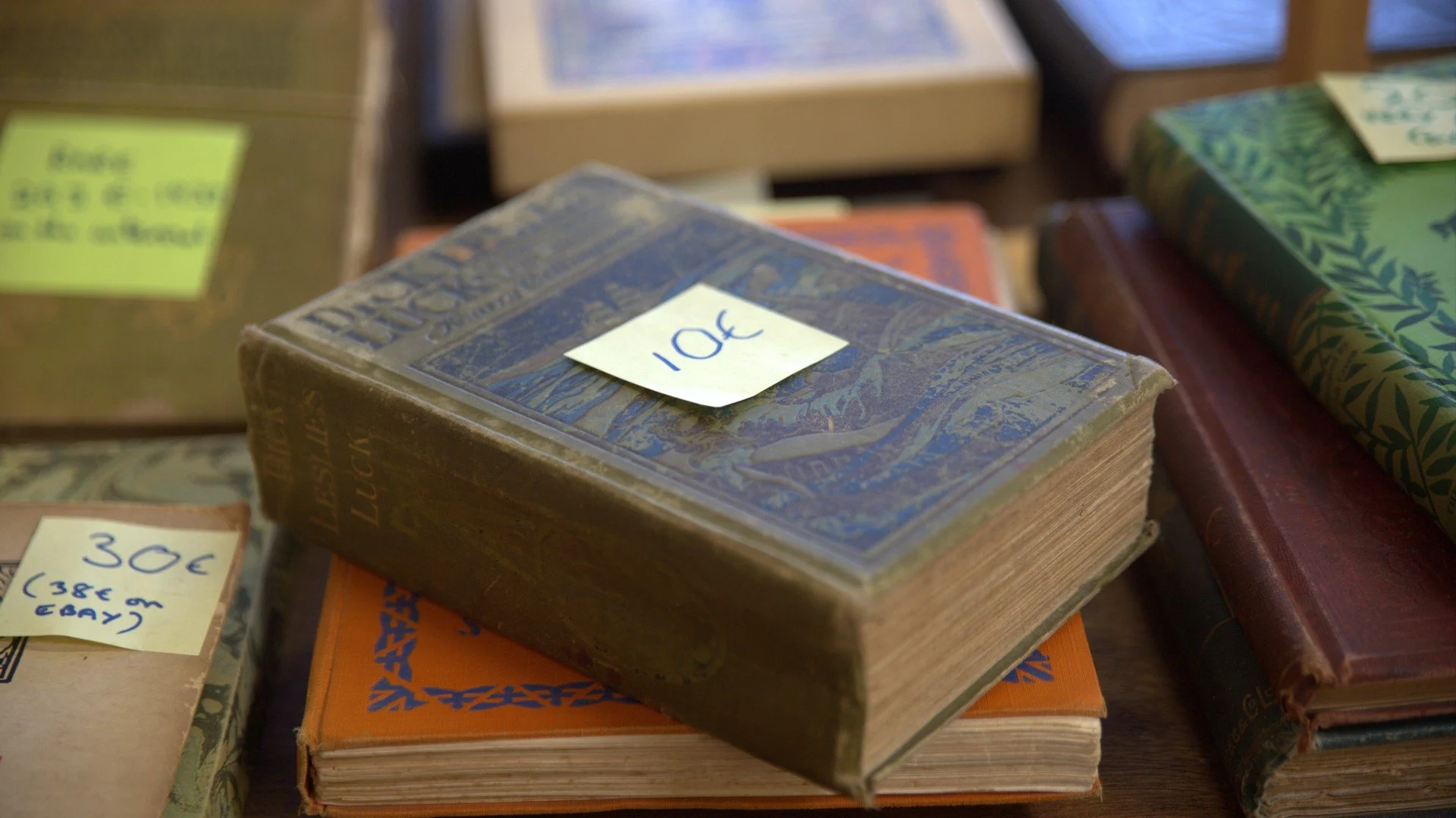 Stack of books at a bookstore with prices marked on sticky notes, including a blue book with a note showing 10 cents and another with 30 euros, among other books with colorful covers.