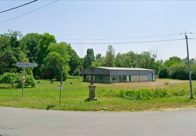 A rural scene with a small building on a grassy area, trees behind, a street sign reading 'Priconrieux', and a decorative cross on a pedestal in the foreground.