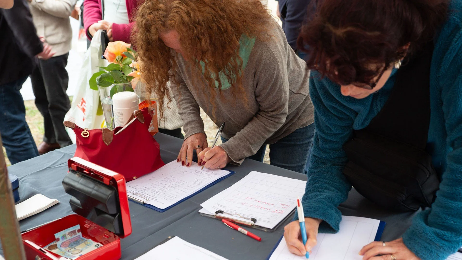 People gathered around a table at an outdoor event, signing documents and writing on paper.