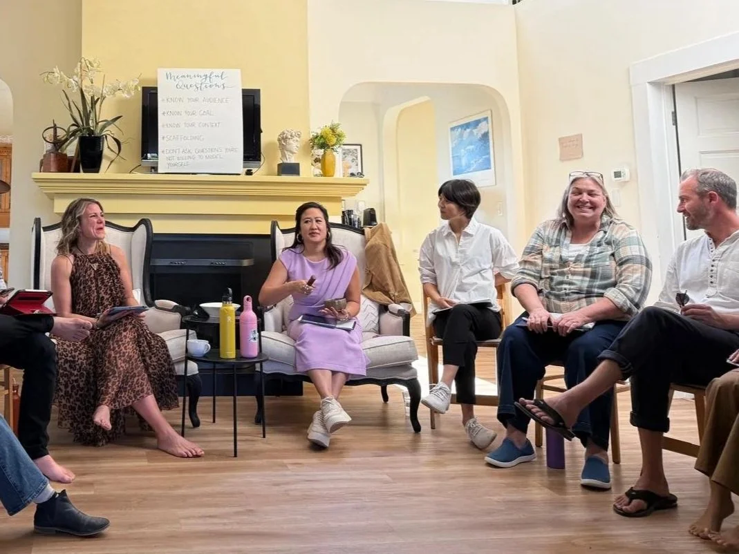 Group of six people sitting and standing in a circle in a living room, engaged in a discussion or activity, with some smiling and others talking.