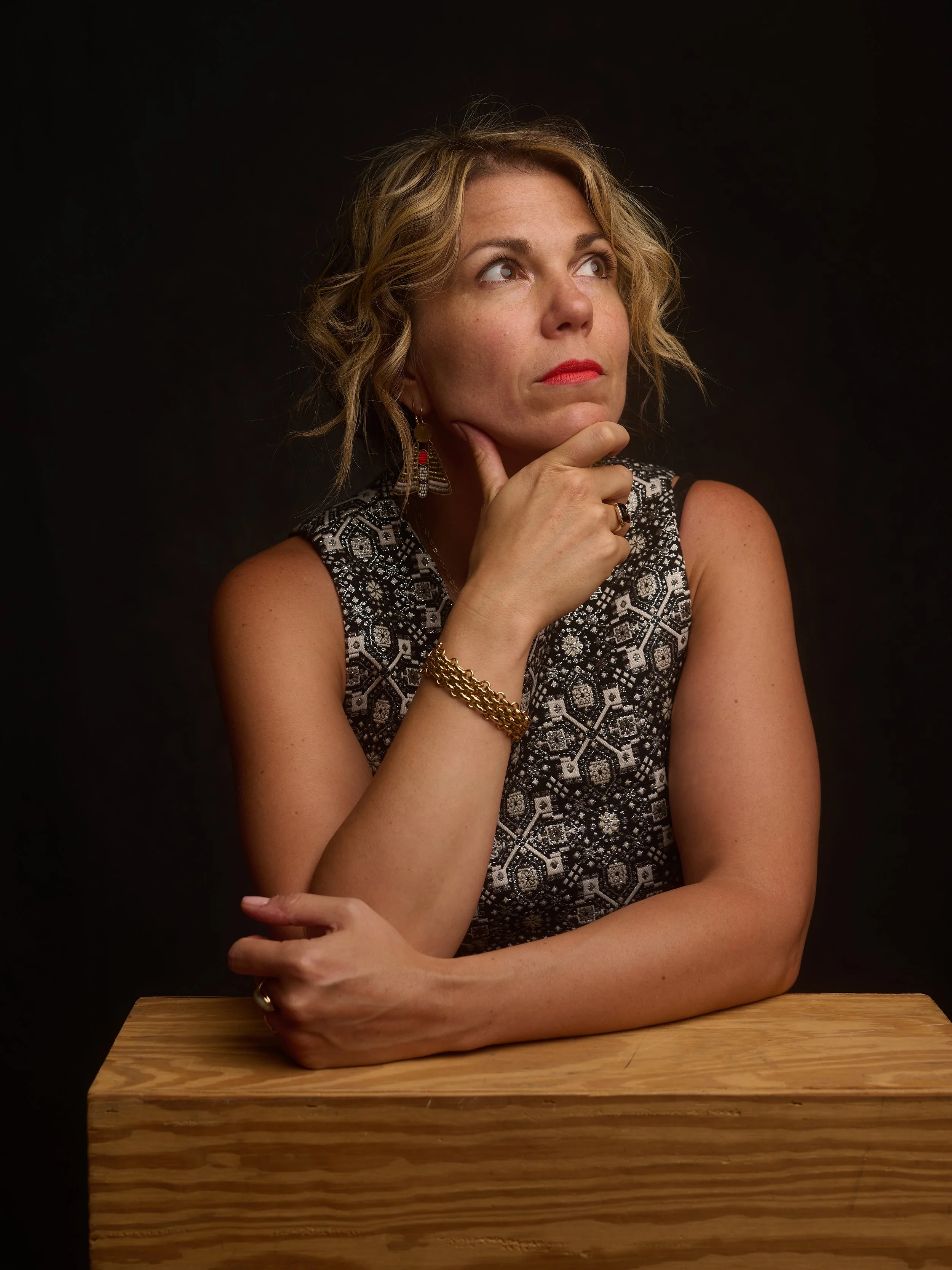 A woman with curly blond hair and red lipstick sits at a wooden table, resting her chin on her hand, with a contemplative expression, against a dark background.