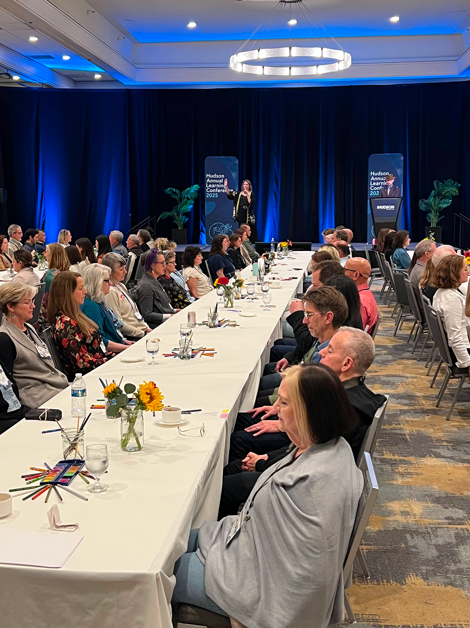 A conference room with attendees seated at long tables covered with white tablecloths, decorated with small vases of sunflowers, water glasses, and colored pencils. A stage at the front has two presenters speaking, with large blue banners displaying 'Hudson Annual Learning Conference 2025.' The room has blue lighting on the back wall, potted plants, and a modern circular chandelier hanging from the ceiling.