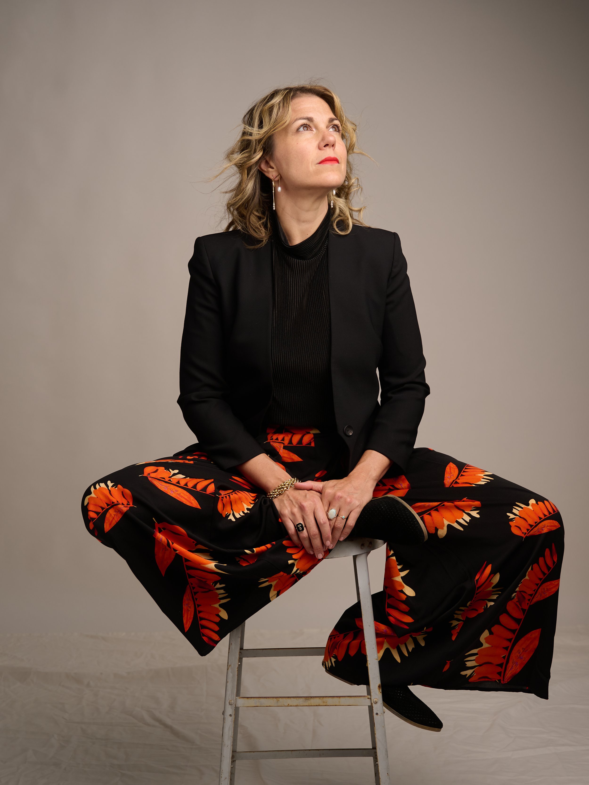 A woman with blonde curly hair, wearing a black blazer, black top, and butterfly patterned orange and black pants, sitting on a white folding chair against a neutral background, looking upwards.