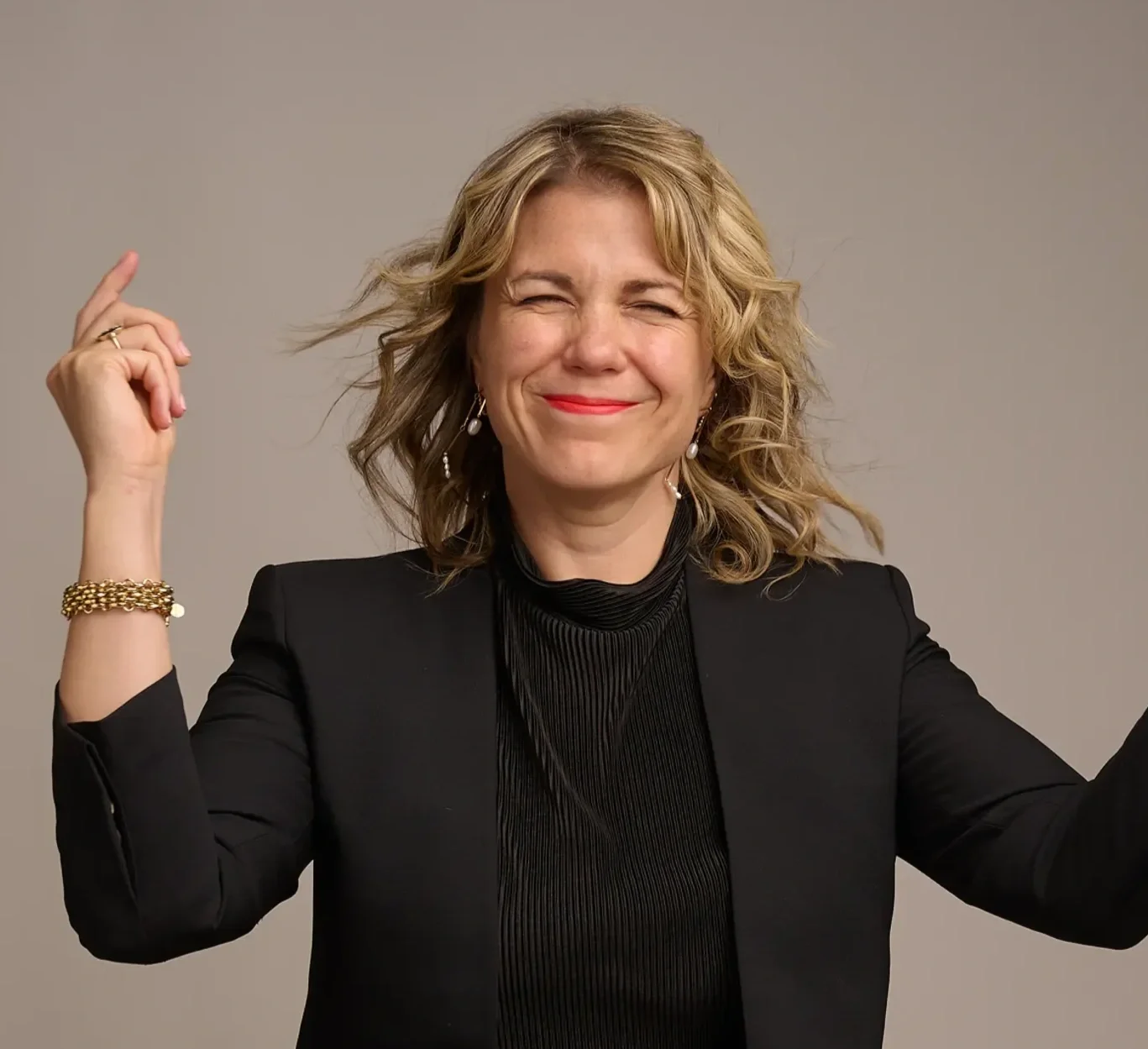 A woman with blonde curly hair smiling and pointing upward, wearing a black blazer and jewelry, against a plain gray background.