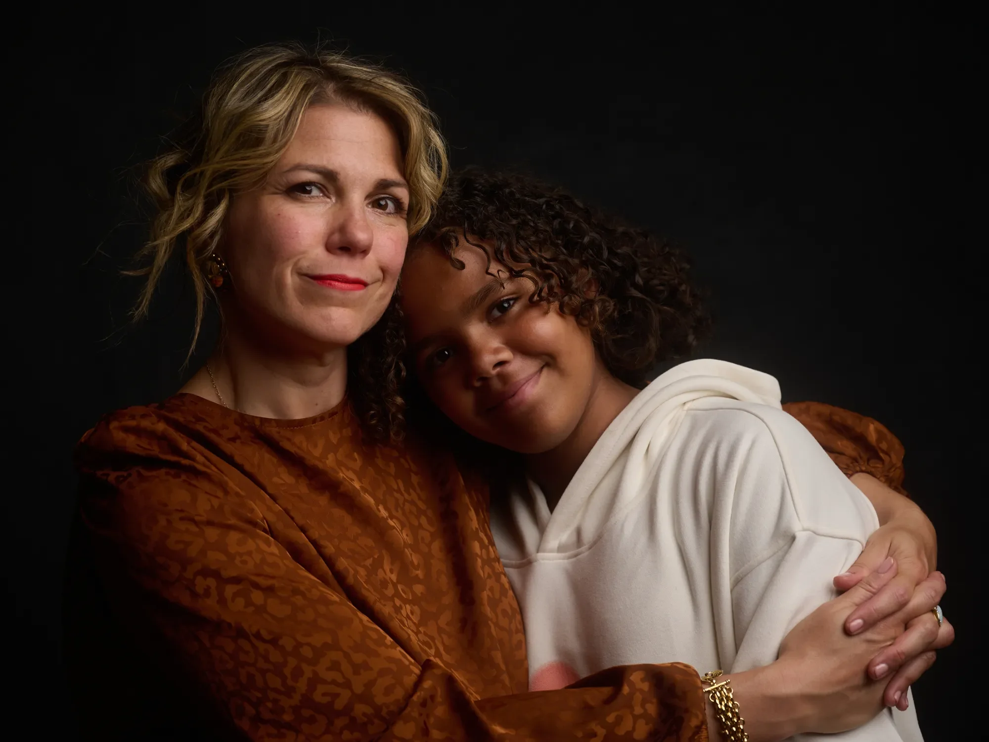 A woman and a young girl sharing a hug against a black background.
