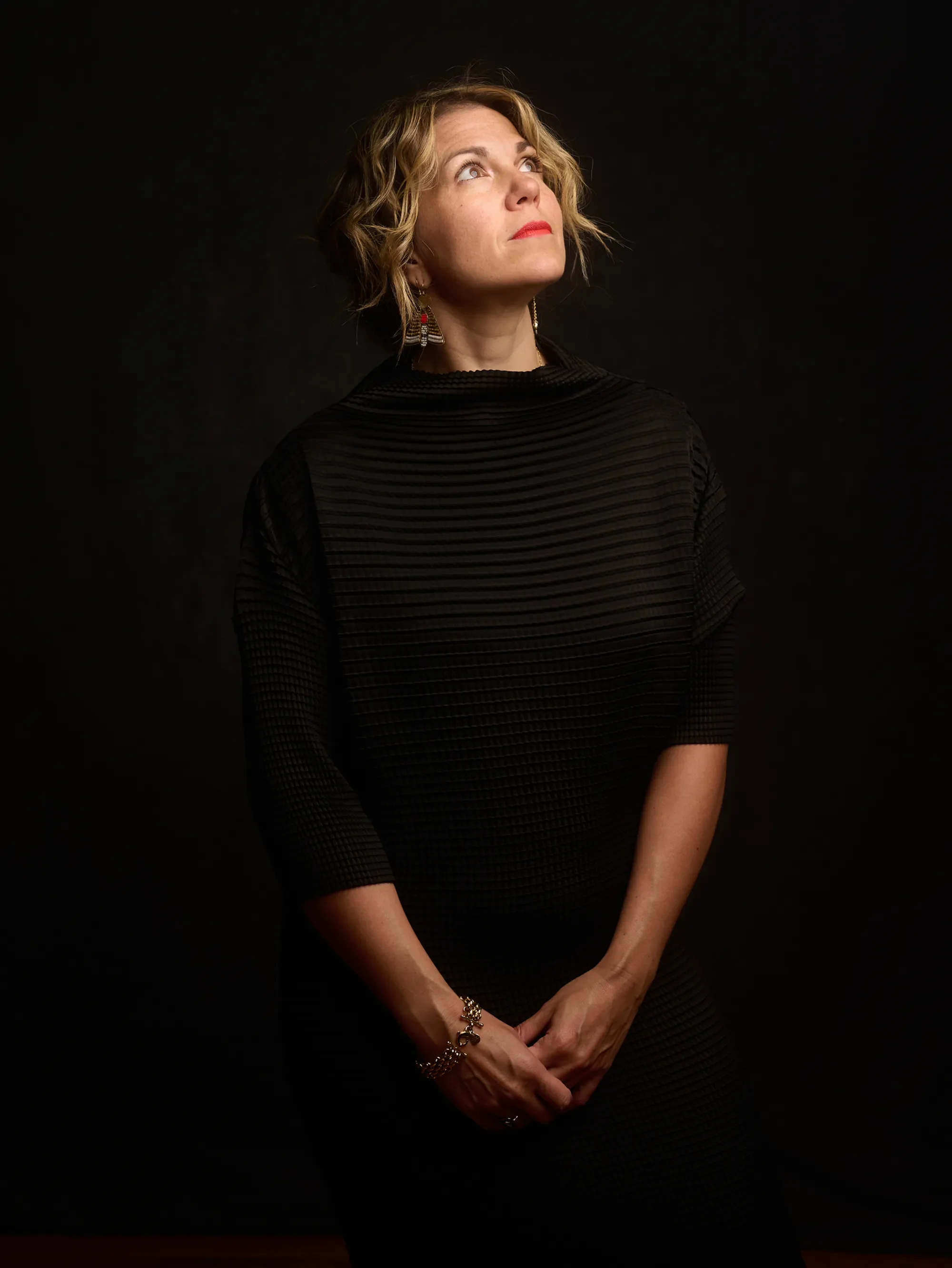 A woman with short curly blonde hair wearing a black top with textured stripes, red lipstick, and large dangling earrings, standing against a dark background and looking upward.