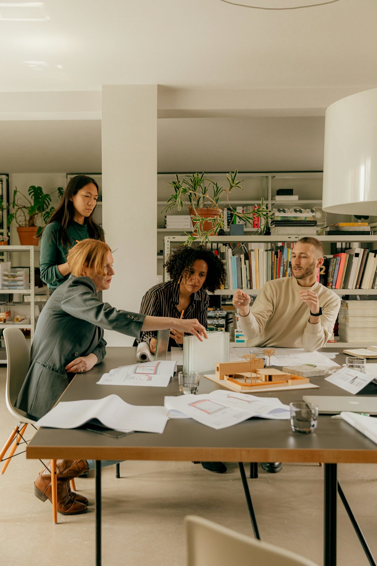 A group of five diverse individuals engaged in a discussion around a table in an office or meeting room, with bookshelves and plants in the background.