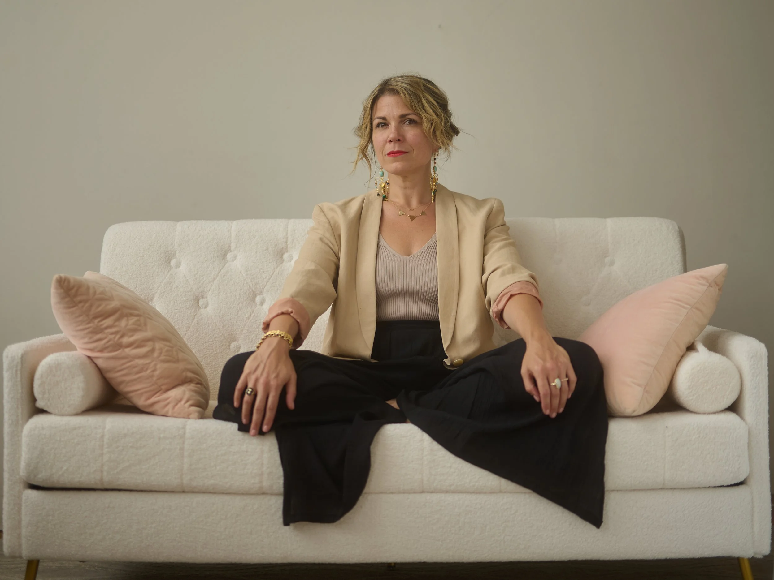 A woman sitting on a cream-colored sofa with pink cushions, wearing a beige blazer, a light top, and black pants. She has short wavy blonde hair, colorful earrings, a necklace, and rings, and is looking at the camera with a slight smile.