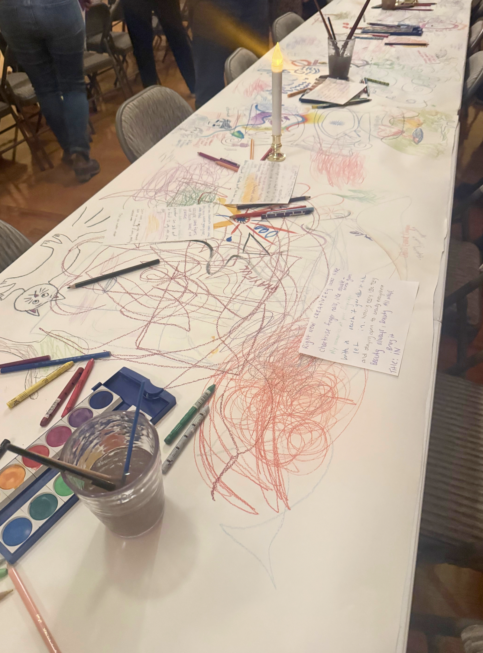 A long table covered with colorful scribbles and drawings made with various markers and crayons in a creative art activity. There are paint palettes, brushes, and notes on the table, with some people standing around in the background.