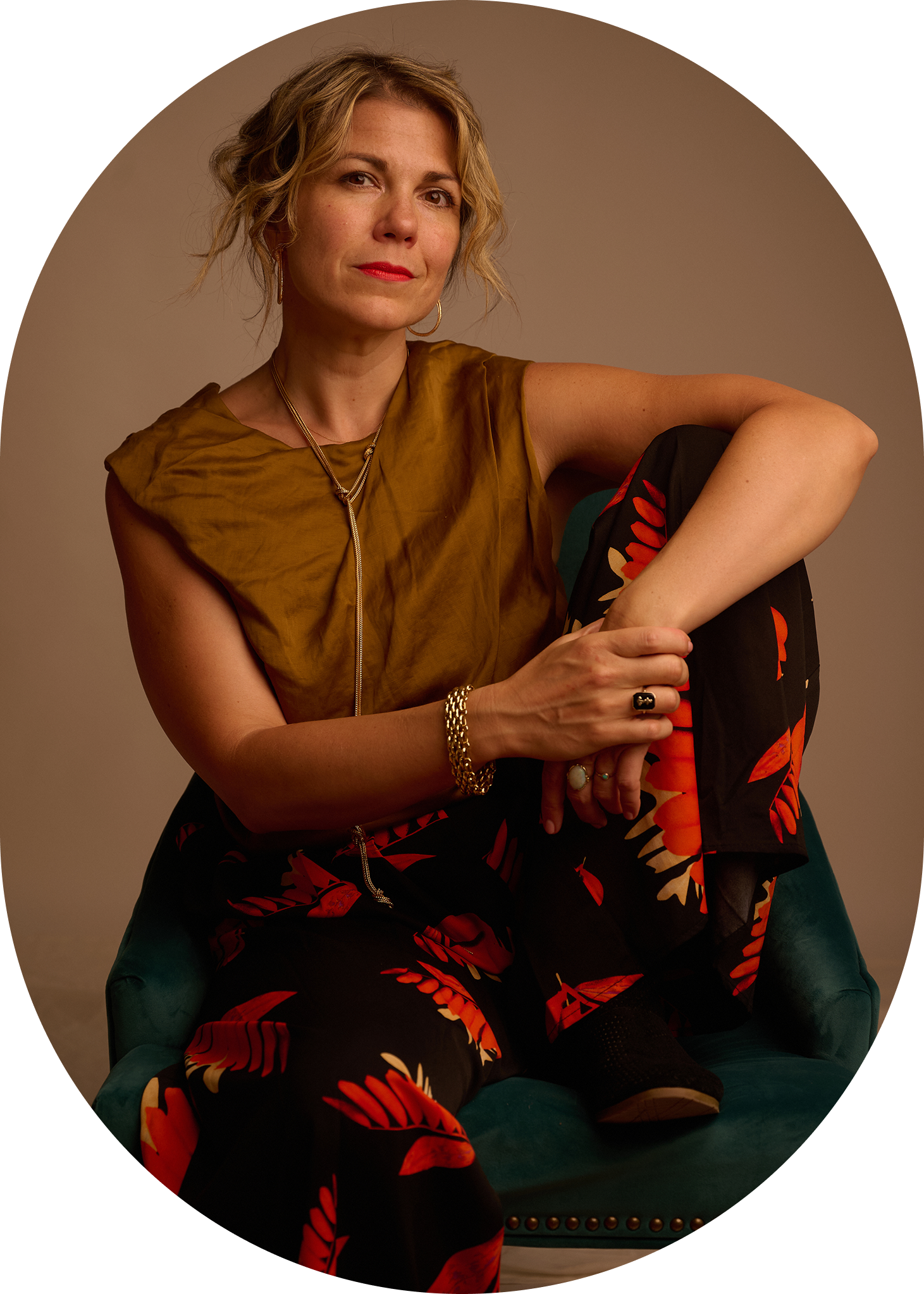 A woman with short, wavy blonde hair and red lipstick is sitting in a green chair with brass nailhead trim. She is wearing a sleeveless, gold-colored top and black pants with a red and orange leaf pattern. She has gold jewelry, including hoop earrings, necklaces, bracelets, and rings, and is looking at the camera with a confident expression. She is sitting with her left arm resting on her knee and her right hand holding her left ankle.