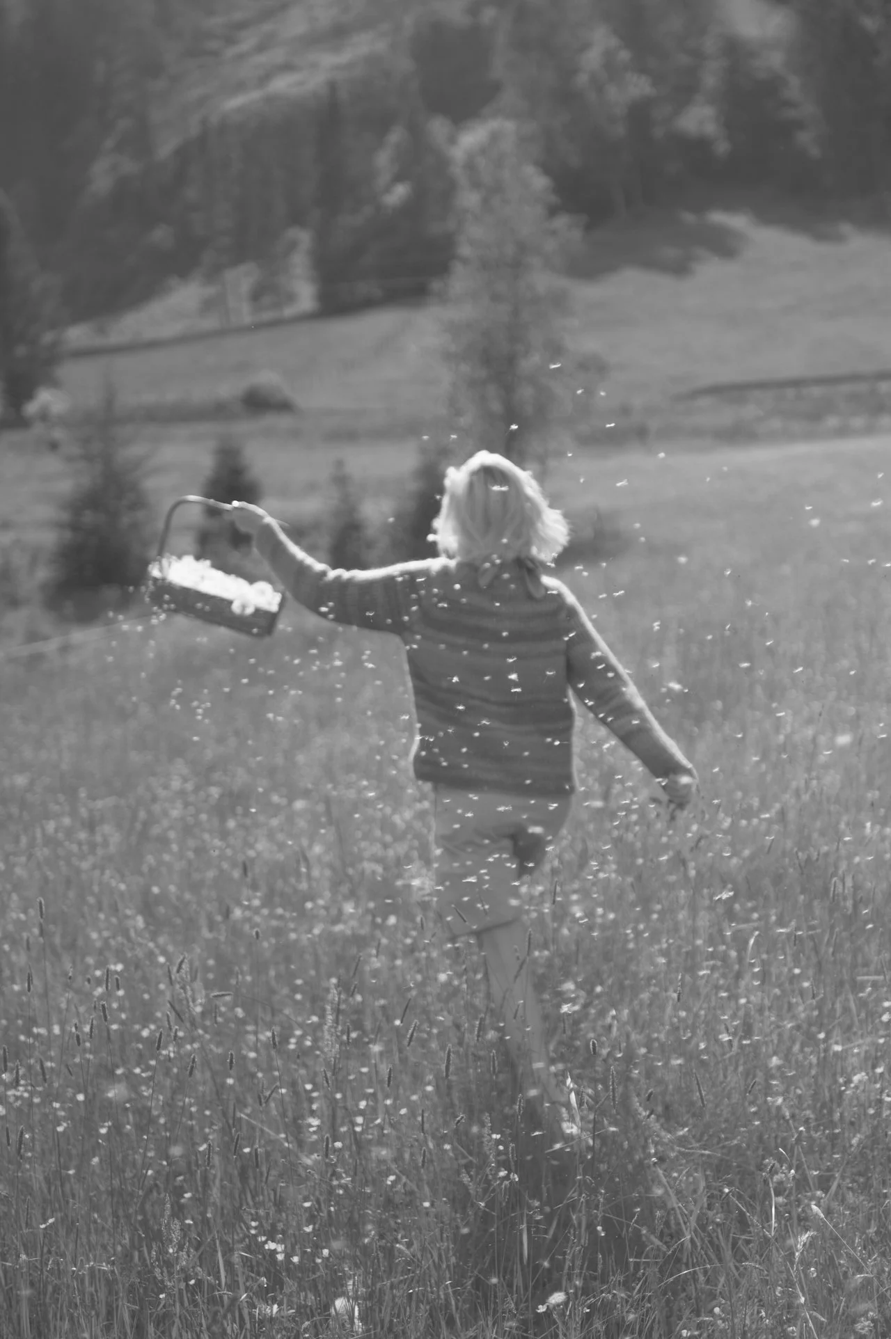 A person with shoulder-length hair, wearing a striped sweater and pants, running through a meadow with tall grass and wildflowers, holding a flower basket, with trees and hills in the background.
