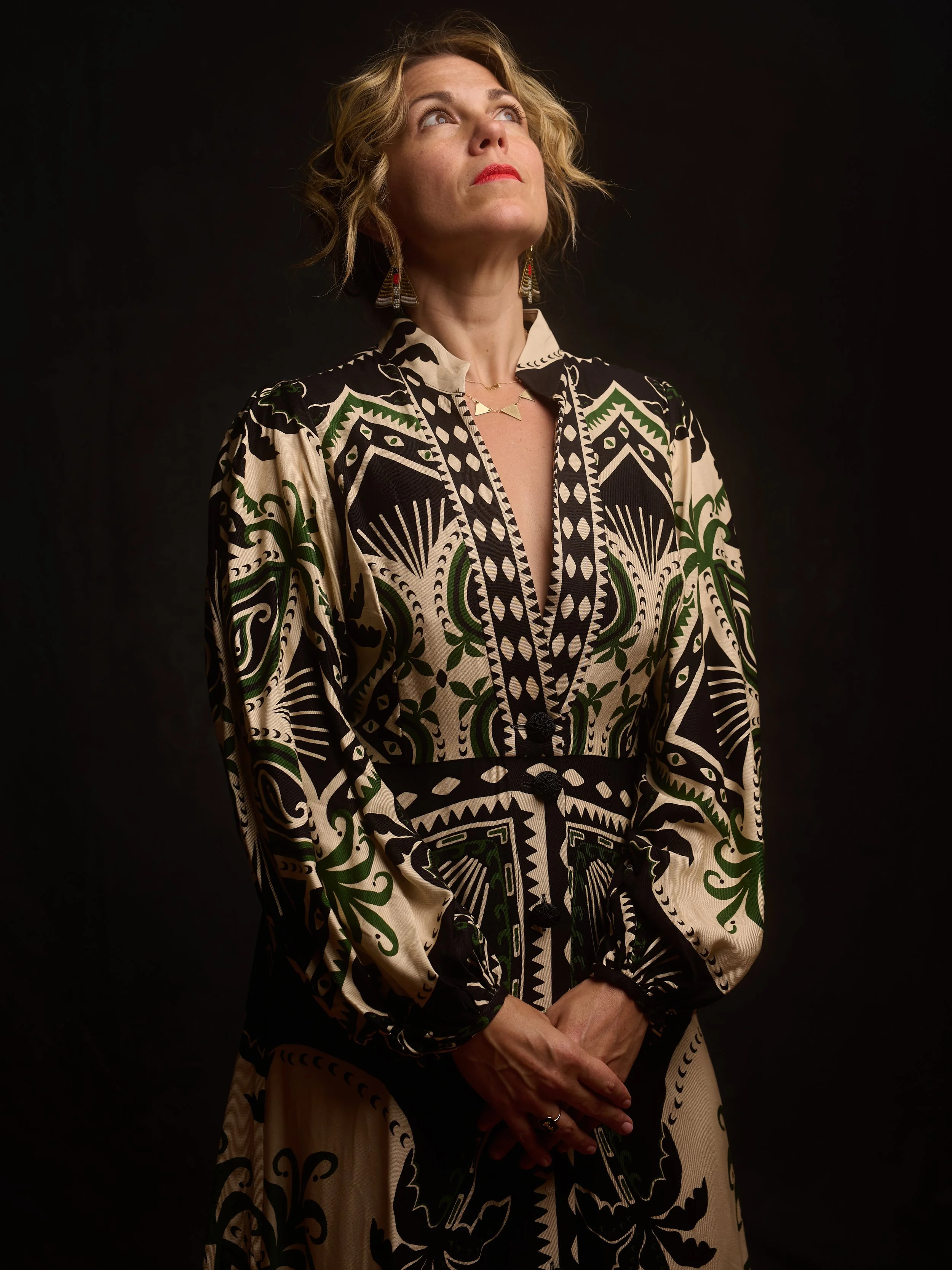 A woman with short curly blonde hair wearing a patterned black, white, and green dress, earrings, and a layered necklace, looking upwards with a dark background.