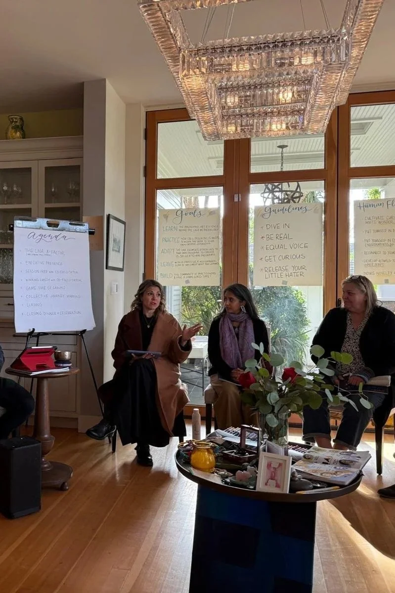 Three women are seated in a living room with a large, modern chandelier hanging above them. They are engaged in a discussion. Behind them are large windows with handwritten notes on paper posters. A table in front of them has flowers, decor, and magazines.