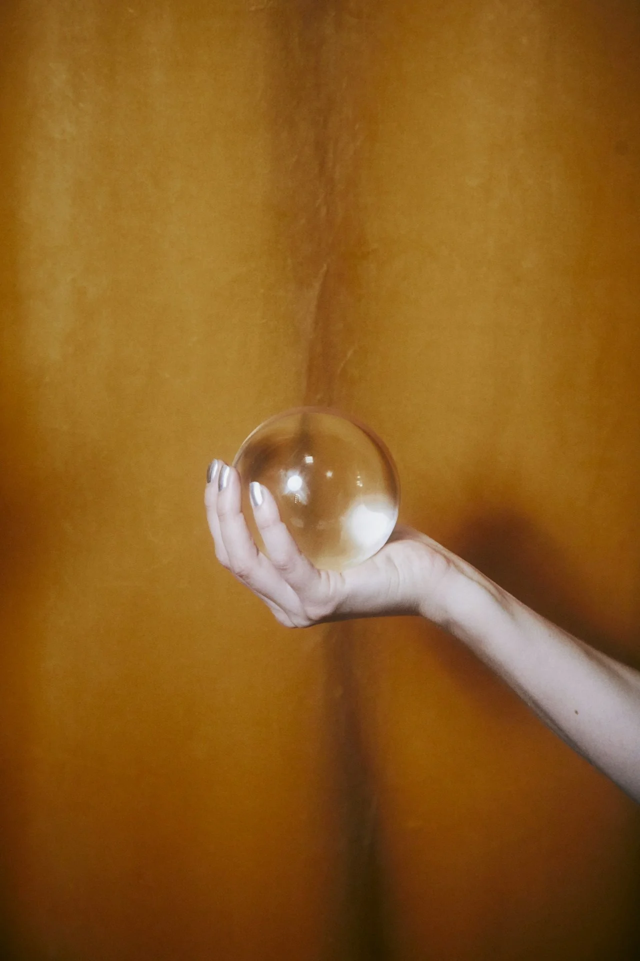A hand with manicured nails holding a clear glass sphere against a warm orange-brown background.