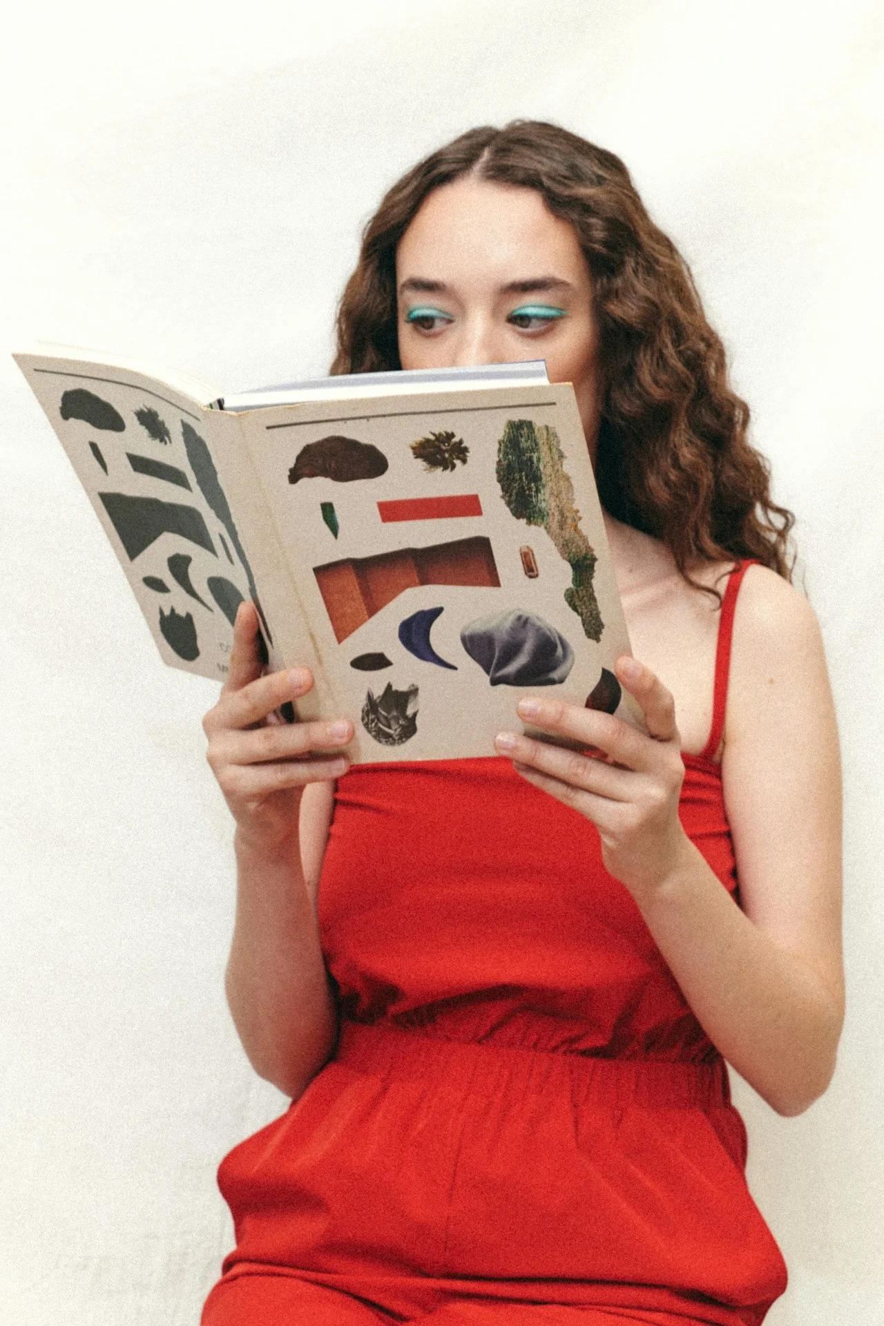 A woman with curly brown hair and teal eye makeup reading a book with various nature and animal illustrations on the cover, wearing a red sleeveless top.