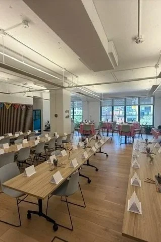 Empty modern event space or conference room with tables set for a gathering, decorated with folded napkins and place cards, featuring large windows and minimalistic decor.