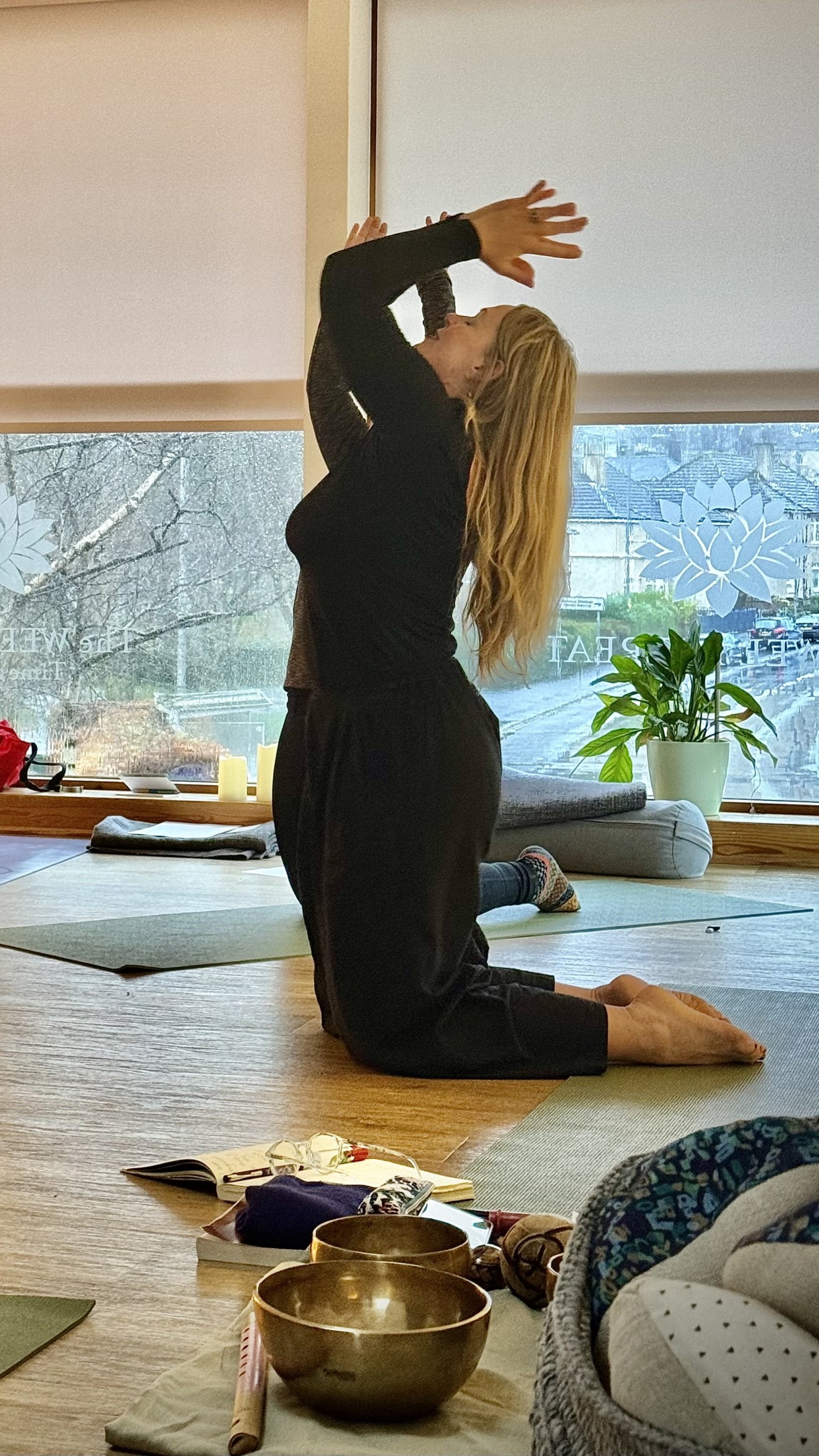 Two women engaging in a yoga or meditation practice inside a room with large windows and a scenic outdoor view. One woman is kneeling and reaching upward with her hands, while the other is kneeling behind her, supporting her.