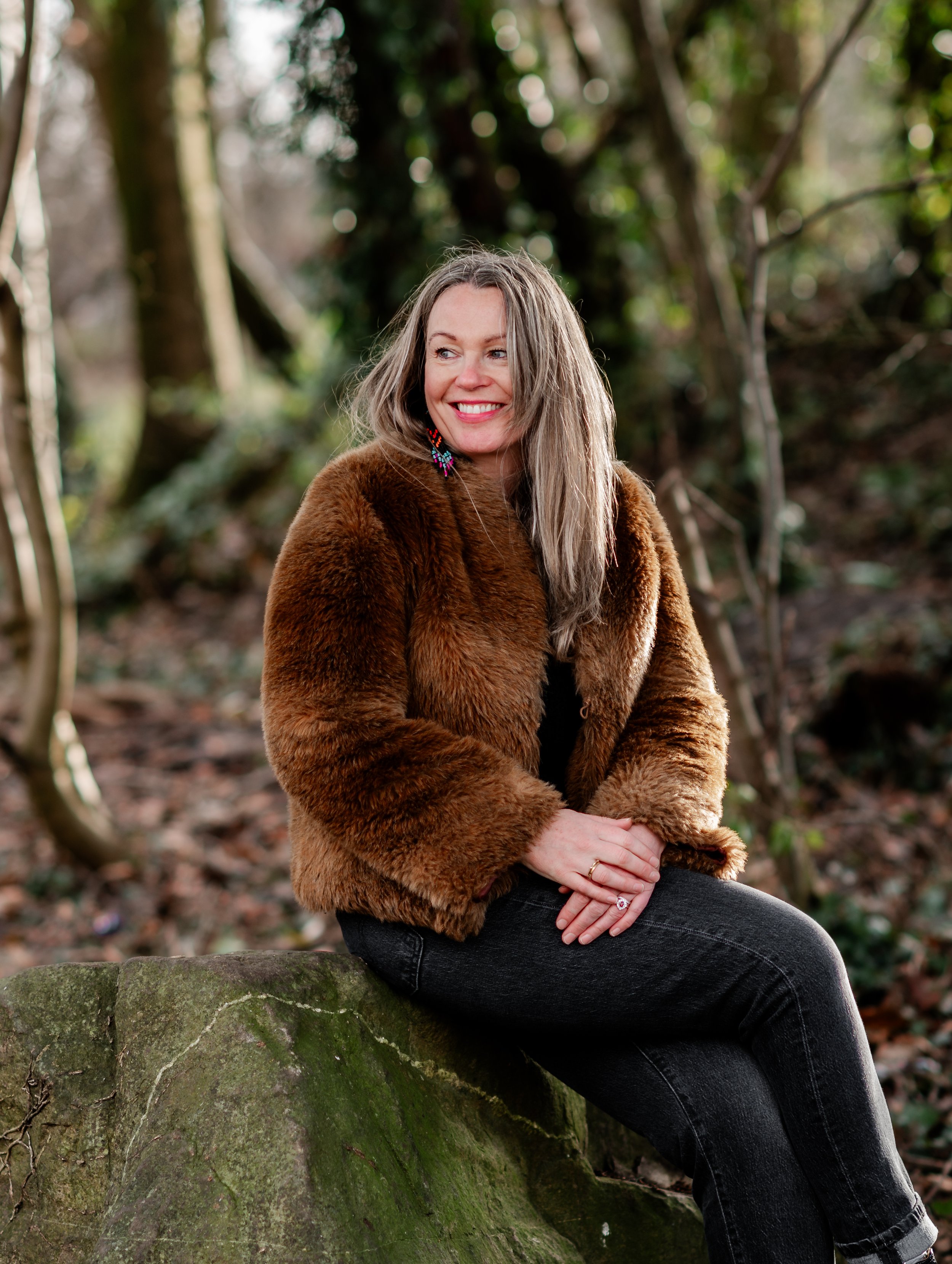 A woman with gray hair, wearing a brown fur coat, sitting on a moss-covered rock in a wooded area, smiling and looking to her right.