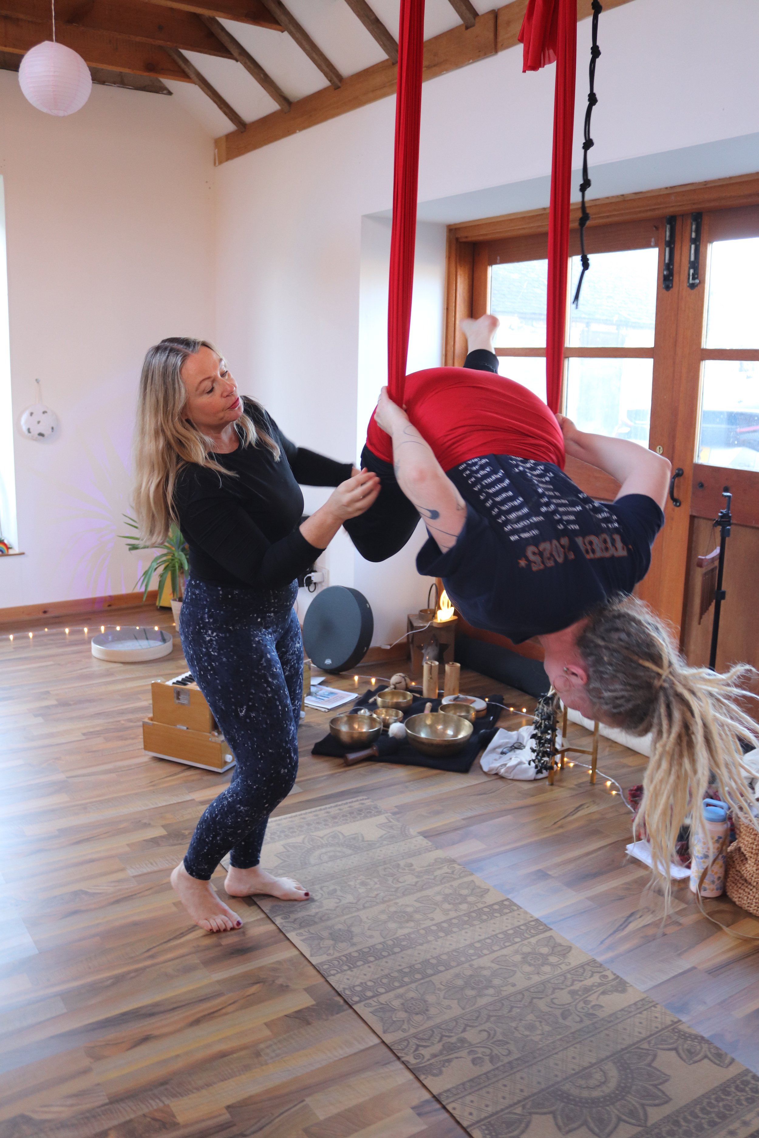 A woman stands on a rug playing a singing bowl while another person hangs upside down from red aerial silk fabric in a cozy wooden room with large windows.