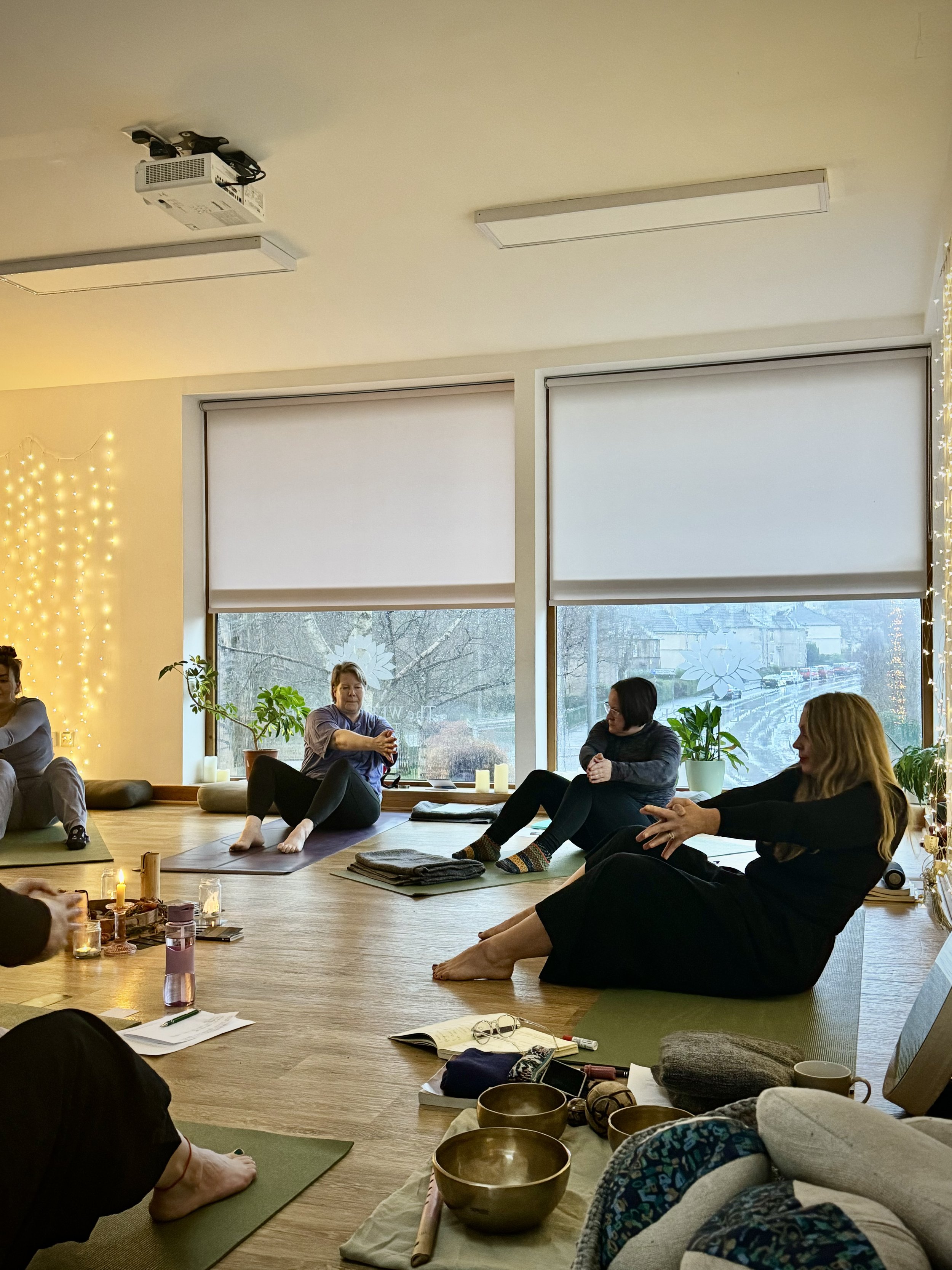People participating in a group yoga or meditation class indoors, sitting on mats with their eyes closed and hands resting on their legs, with candles and singing bowls on the floor, large windows with view outside, and soft lighting.