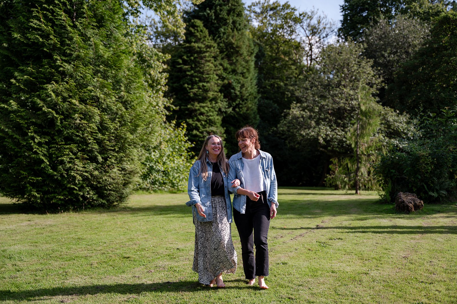 Two women walking and talking in a park with green trees and grass, smiling and enjoying each other's company.